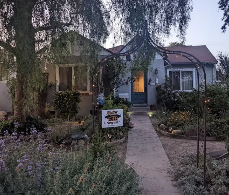 Front yard of a small home with a native plant garden, a lighted metal archway over the walkway, and a “Beautiful Yard 2022 Congrats!” sign near the entrance at dusk.